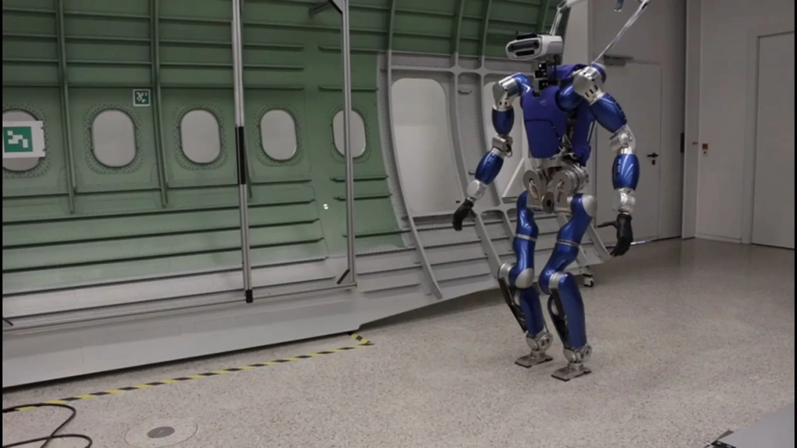 Blue humanoid robot walking in a test hall in front of an aircraft fuselage mock-up